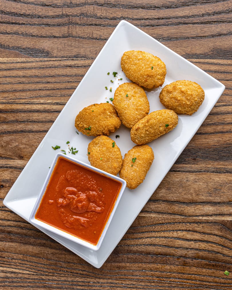 Crispy golden-brown croquettes with green herbs, served with a side of red marinara sauce on a white plate.
