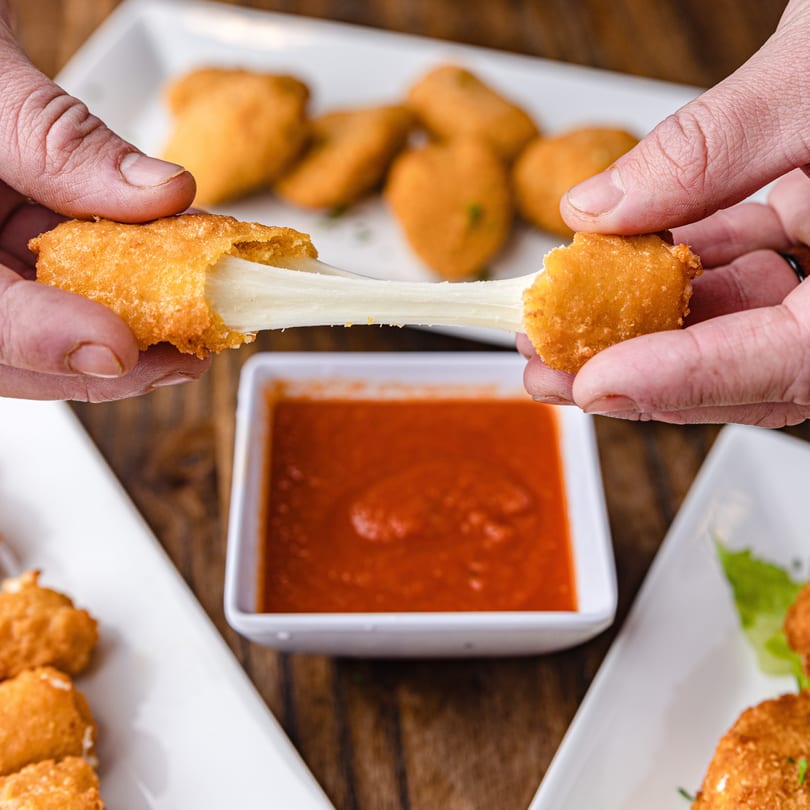 Hands pulling apart a fried mozzarella stick, revealing stretchy cheese, with marinara sauce nearby.