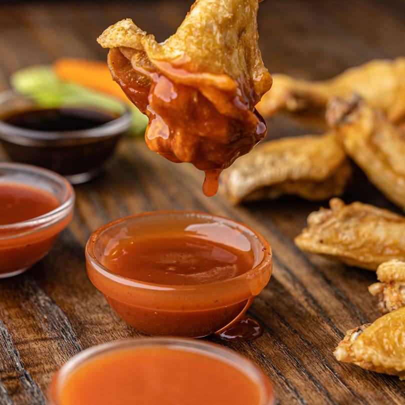 Crispy chicken wing being dipped into a bowl of orange sauce, with other sauces and wings in the background.