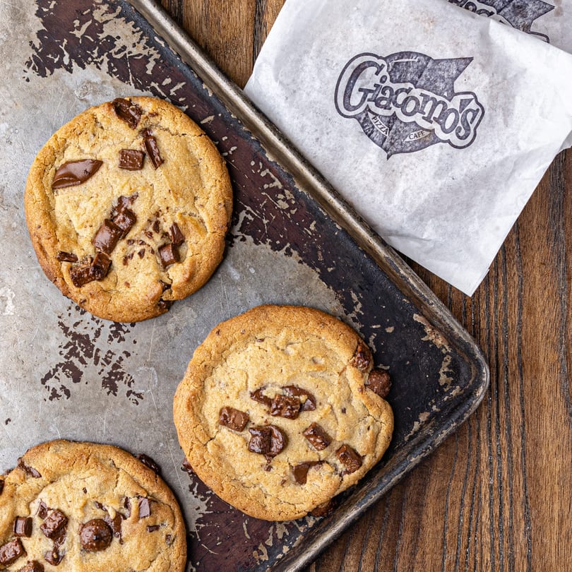 Three chocolate chip cookies on a metal tray next to a Giacomo's Pizza Cafe branded paper.